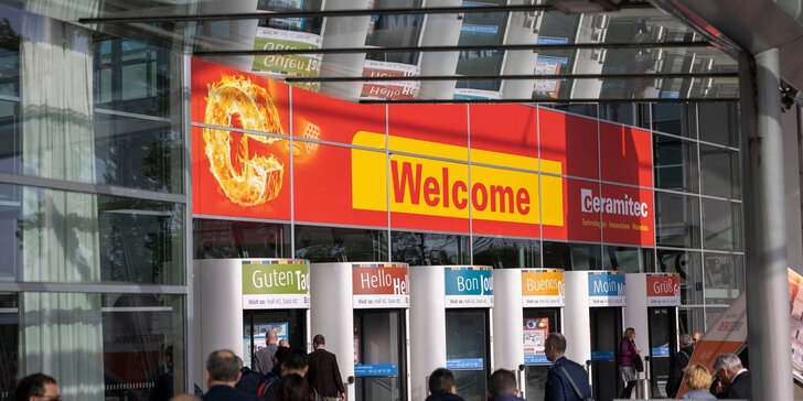 Several revolving doors leading to the entrance hall of the ceramitec trade fair, with a large red welcome sign above them and numerous people entering the building.