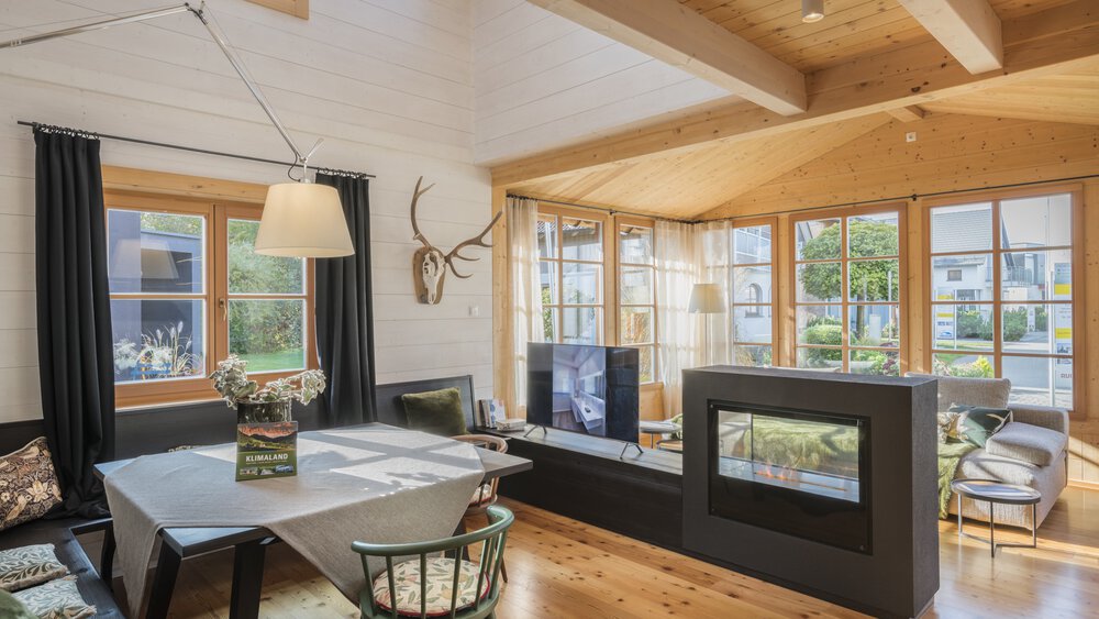 Ein gemütliches Wohnzimmer mit Holzbalken, Esstisch, Kamin, an der Wand montiertem Hirschgeweih und großen Fenstern.