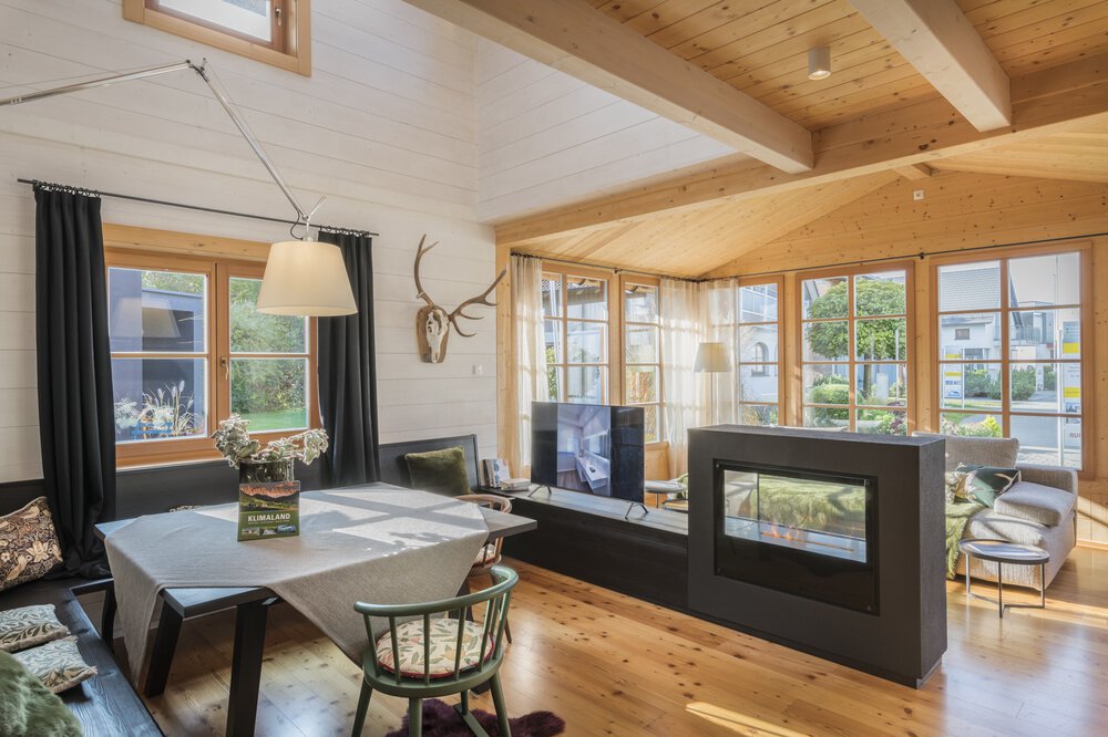 Ein gemütliches Wohnzimmer mit Holzbalken, Esstisch, Kamin, an der Wand montiertem Hirschgeweih und großen Fenstern.