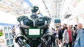 A futuristic, humanoid robot with green glowing elements stands in an exhibition hall in front of a crowd of people who are filming it.