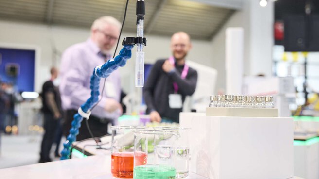 Close-up of a laboratory device. A kind of pipette hovers over measuring beakers with red and green liquid