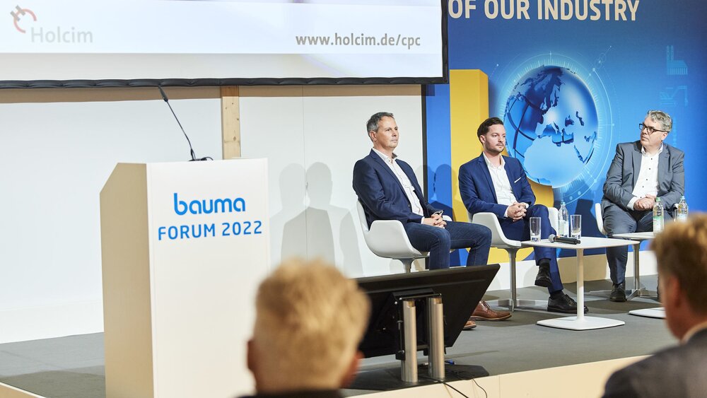 Three men in suits sit on a stage at the bauma Forum 2022, a screen can be seen in the background.