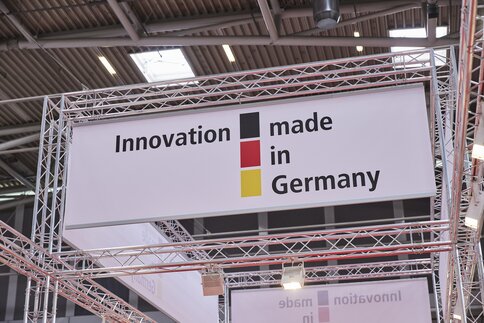 An advertising banner with the words “Innovation made in Germany” and the colours of the German flag hangs from a crossbeam in an exhibition hall.