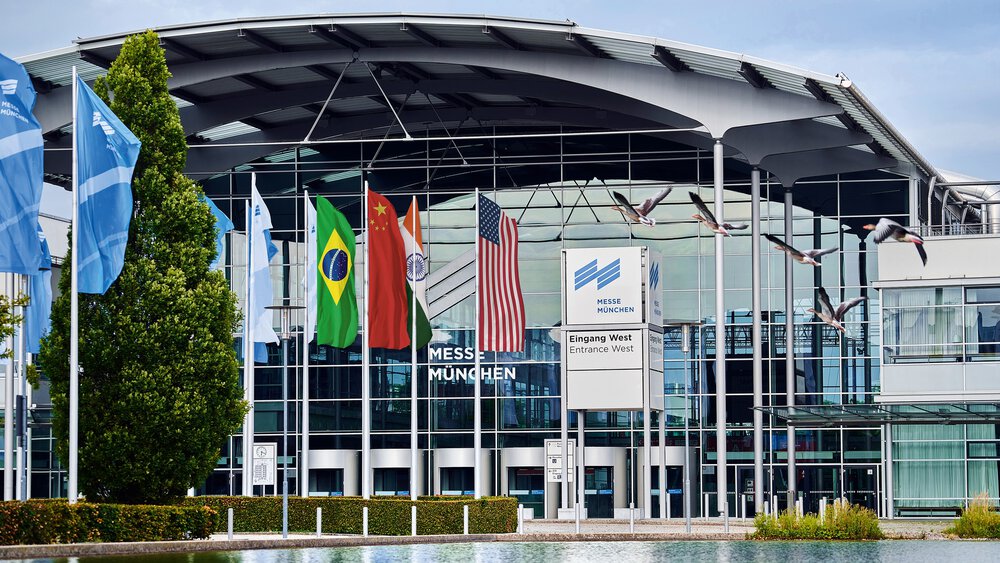 Gebäude vom Eingang West des Messe München Geländes mit Messesee und mehreren Flaggen an Fahnenmasten, z.B. USA, China & Indien.