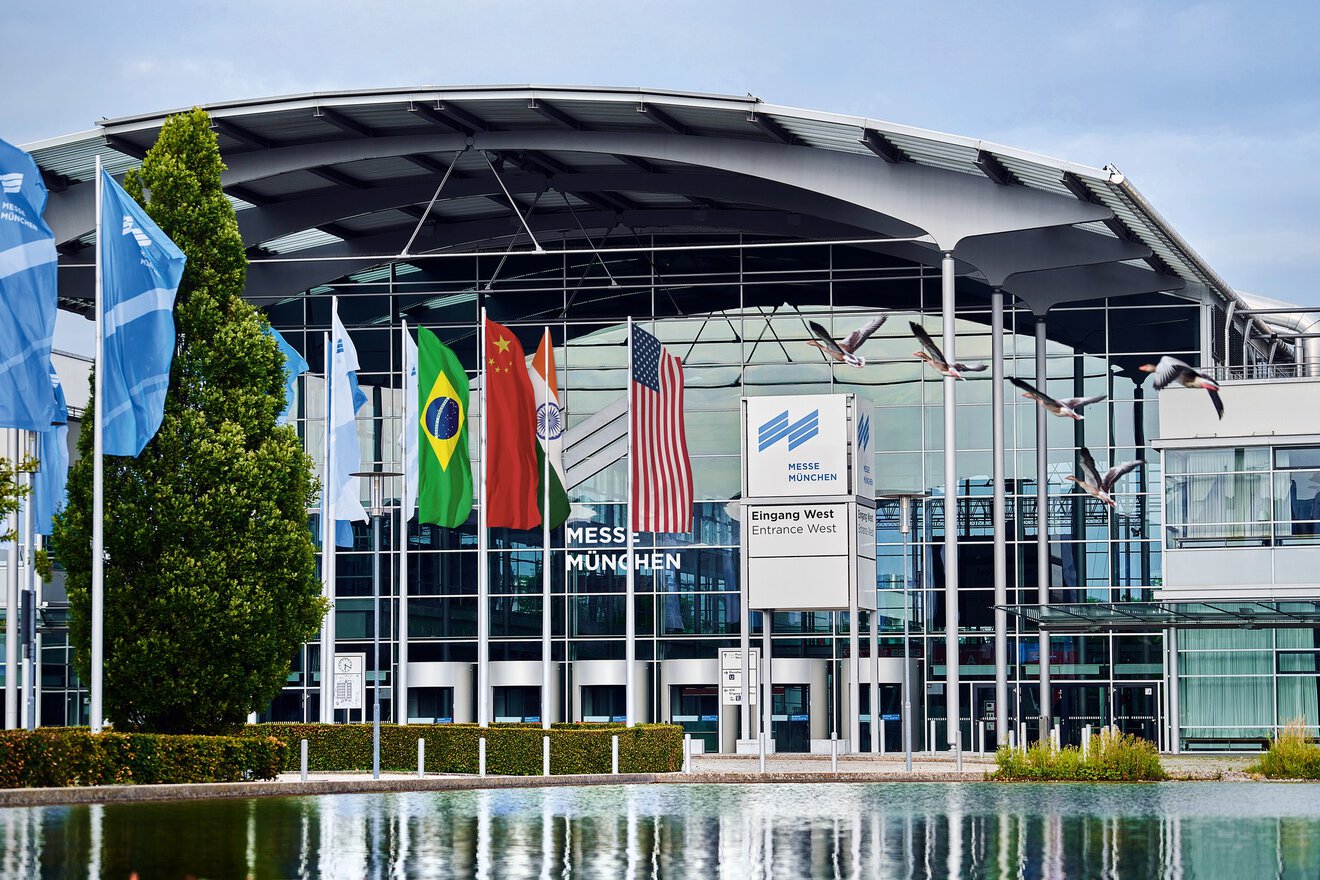 Gebäude vom Eingang West des Messe München Geländes mit Messesee und mehreren Flaggen an Fahnenmasten, z.B. USA, China & Indien.