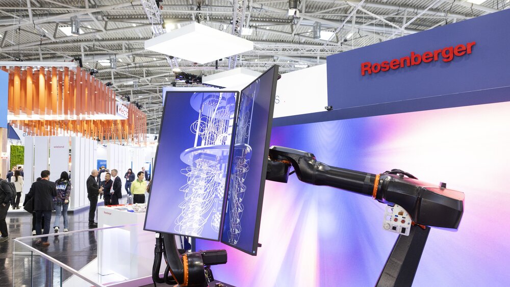 Robot arm with touchscreen display at an exhibition stand, with other exhibition stands and visitors in the background.