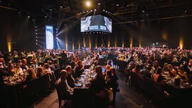 A well-attended ballroom with lots of tables, spotlights on the ceiling and a stage in the background with 