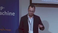 Ein Mann mit Brille und Blazer hält einen Vortrag. Er steht neben einer Leinwand und hinter ihm ist ein blaues Banner zu sehen.