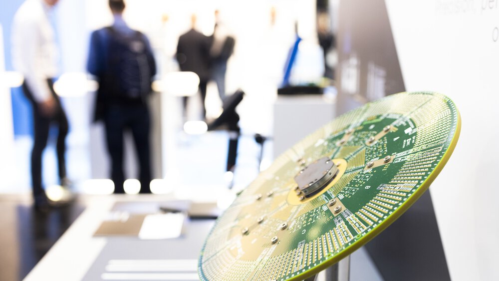 Nahaufnahme einer ausgestellten grünen Leiterplatte mit unscharfen Personen in Geschäftskleidung im Hintergrund.