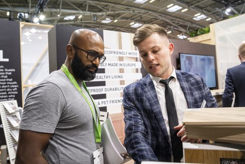 Zwei Männer betrachten etwas an einem Messestand auf der BAU. Hinter ihnen sind eine bedruckte Rückwand, Prospekte und ein Bildschirm zu sehen.