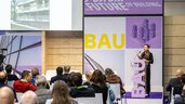A speaker stands at the podium in the BAU forum and gives a lecture to the audience. The decorative elements on the stage are purple and yellow.