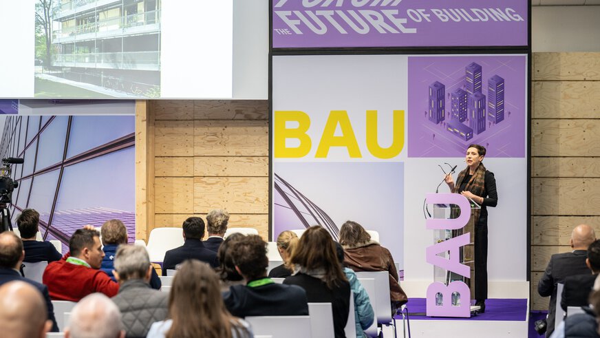 Im Forum der BAU steht eine Rednerin am Podium und hält einen Vortrag vor Publikum. Die Dekoelemente der Bühne sind in lila und gelb gehalten.