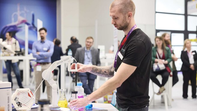 A person stands at a stand and interacts with a white technical device. Other trade fair visitors can be seen in the background