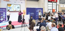 Zwei Personen sprechen auf einer Bühne mit Mikrofonen vor Publikum und Kameras bei einer Veranstaltung von Odense Robotics.