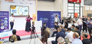 Two people speak with microphones on stage in front of an audience and cameras at an Odense Robotics event.