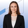 Junge Frau mit schulterlangem braunen Haar, schwarzem Blazer und weißer Spitzenbluse vor modernem, blau-grafischem Hintergrund.