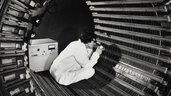 A person in a lab coat sits in a tubular metal structure and inspects the surface with scientific equipment.