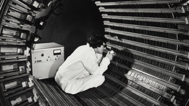 A person in a lab coat sits in a tubular metal structure and inspects the surface with scientific equipment.