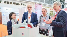 Four trade fair visitors examine a technical exhibit together while one explains something.