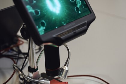 Ein digitales Mikroskop auf einem Tisch. Es ist eingeschaltet und zeigt eine Nahaufnahme einer Platine.