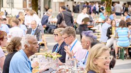 Menschen sitzen auf Bierbänken im Freien und unterhalten sich bei einer gut besuchten Veranstaltung.