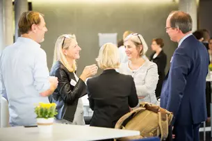 Fünf Personen stehen und unterhalten sich bei einer zwanglosen Veranstaltung im Innenbereich, mit Kaffeetassen und einer kleinen Pflanze auf dem Tisch.