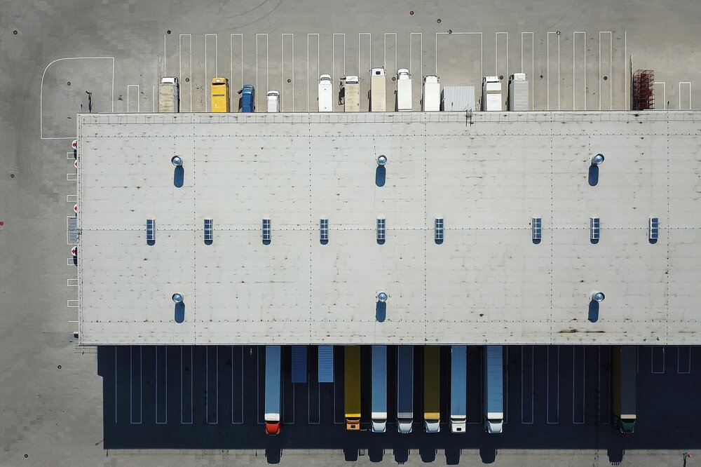 A view from above of a large grey warehouse roof with numerous delivery bays for lorries and lorries parked in reverse alongside the warehouse.