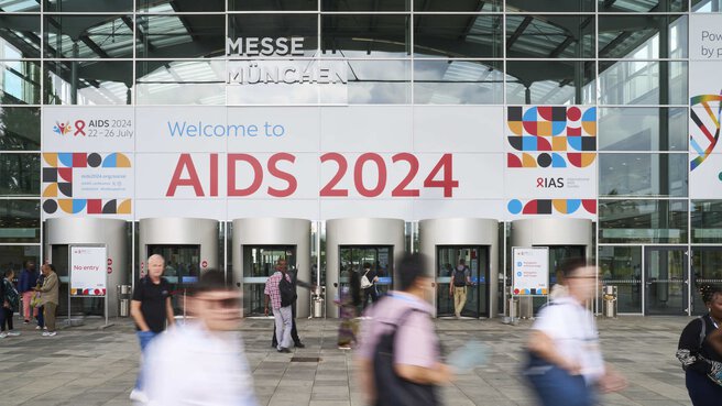 Menschen gehen an einem Eingang mit einem großen AIDS 2024-Banner auf dem Gelände der Messe München vorbei.