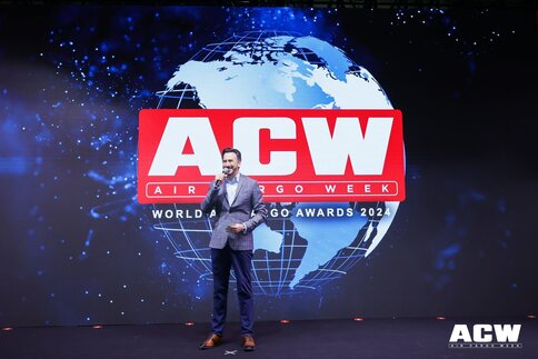 A man in a suit speaks with a microphone in front of a globe backdrop on the stage. The red logo of the 