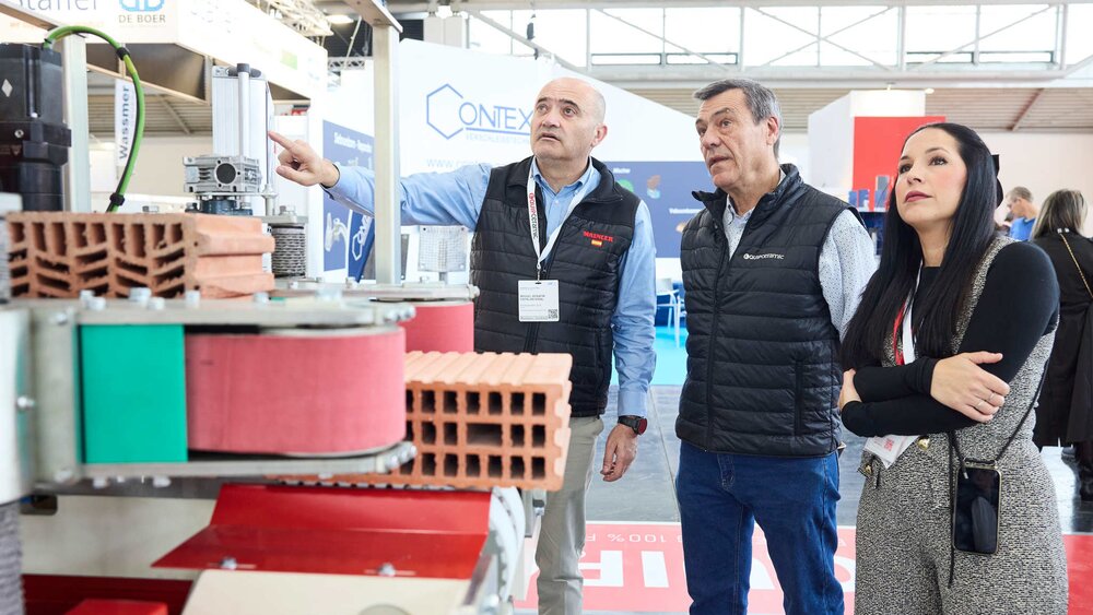 Three trade fair visitors observe a brick-processing machine while one explains something.