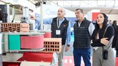 Three trade fair visitors observe a brick-processing machine while one explains something.