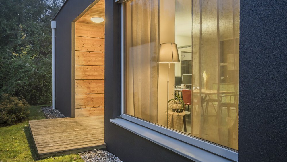 Modernes Haus mit großen Glasfenstern durch die ein elegantes Wohnzimmer zu sehen ist, warmes Licht scheint aus den Fenstern in die Abenddämmerung.