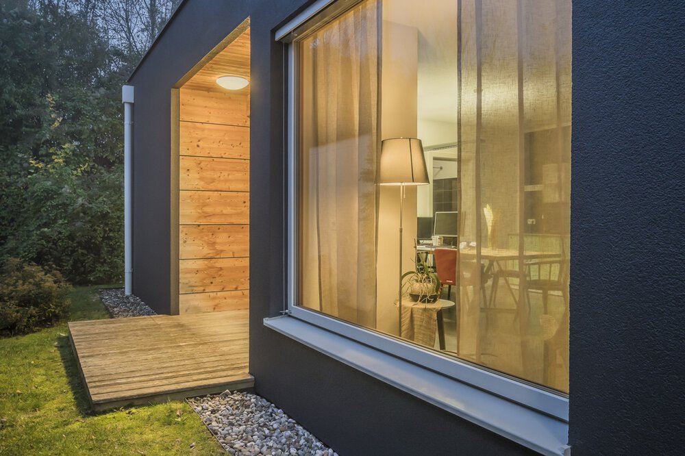 Modernes Haus mit großen Glasfenstern durch die ein elegantes Wohnzimmer zu sehen ist, warmes Licht scheint aus den Fenstern in die Abenddämmerung.