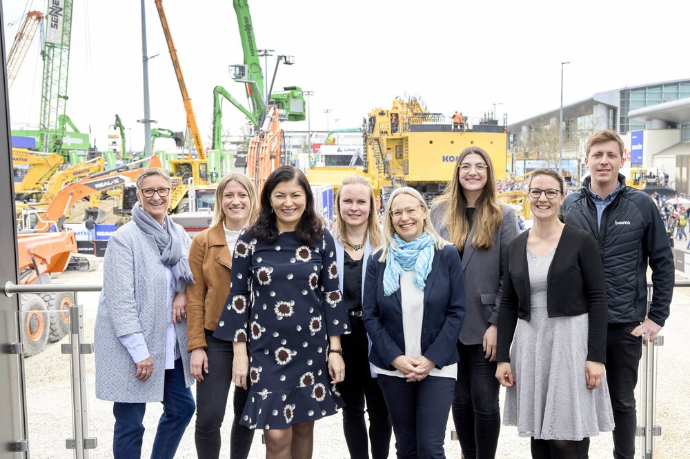 Group of people posing in front of construction machinery at an outdoor trade fair.
