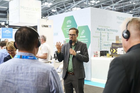 Man in suit speaks with microphone at trade fair, listeners wear headsets