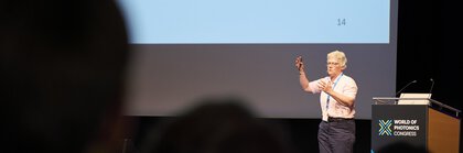 Person giving a presentation on stage at the World of Photonics Congress, with a slide on UV and X-rays in the background.