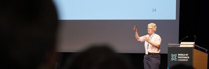 Person giving a presentation on stage at the World of Photonics Congress, with a slide on UV and X-rays in the background.