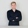 Ein Mann mit kurzen blonden Haaren in einem schwarzen Hemd posiert lächelnd und mit verschränkten Armen.