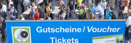 Viele Menschen wartet im Eingangsbereich der INTERFORST auf den Einlass. Im Vordergrund dominiert ein blaues Schild mit Aufschrift 