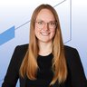 Eine lächelnde Frau mit sehr langen rötlichen Haaren trägt eine Brille, eine Halskette, ein schwarzes Oberteil und einen schwarzen Blazer.