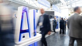 Ein Gang in einer Halle der productronica Messe mit zwei blau erleuchteten Buchstaben, die „AI“ darstellen und mehreren Besuchern, die daran vorbeigehen.