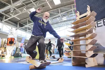 A man balances on a balance board at an exhibition stand. To his right is a stand with other wooden boards.