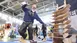 A man balances on a balance board at an exhibition stand. To his right is a stand with other wooden boards.