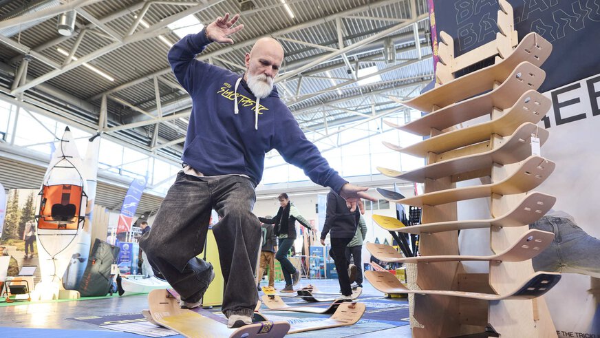 Ein Mann balanciert auf einem Balance-Board an einem Messestand. Rechts neben ihm steht ein Aufsteller mit weiteren Boards aus Holz.