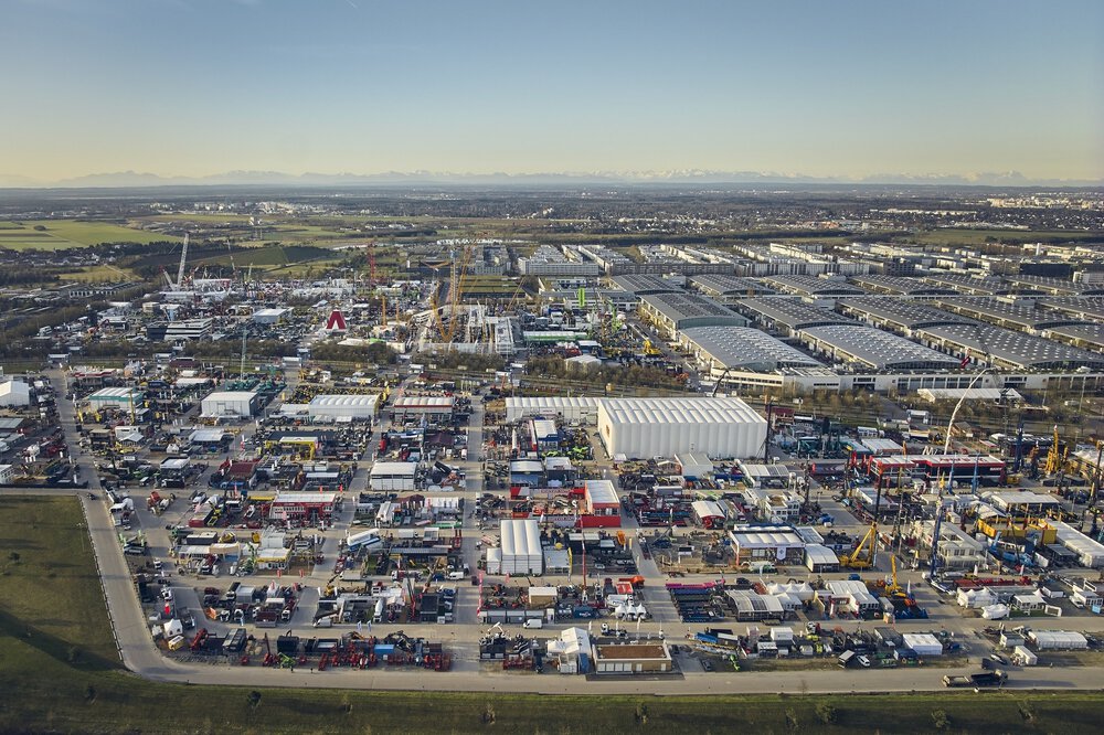 Luftaufnahme eines großen Industriegebiets mit zahlreichen Gebäuden, Fahrzeugen und Geräten. Im Hintergrund sind Berge zu sehen.