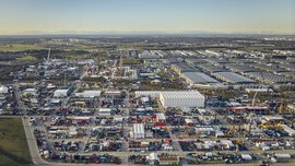 Luftaufnahme eines großen Industriegebiets mit zahlreichen Gebäuden, Fahrzeugen und Geräten. Im Hintergrund sind Berge zu sehen.