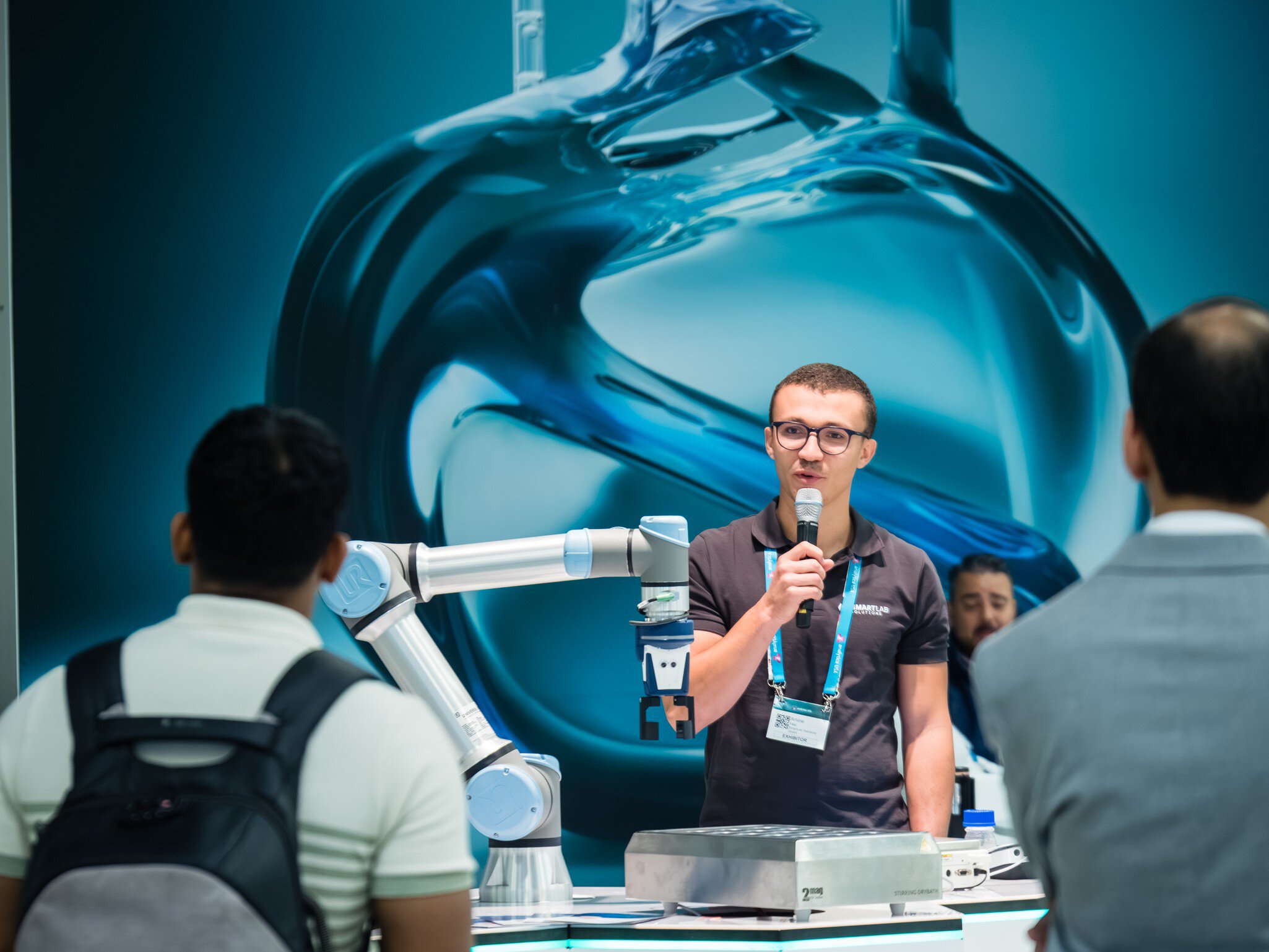 A man speaks into a microphone next to a robotic arm while people watch at an exhibition.