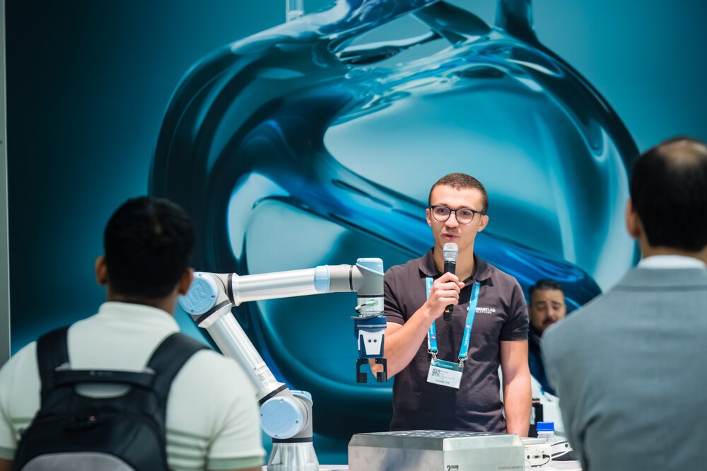 A man speaks into a microphone next to a robotic arm while people watch at an exhibition.