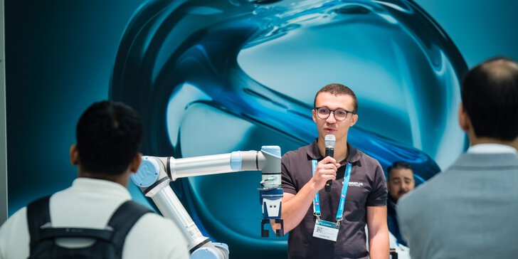A man speaks into a microphone next to a robotic arm while people watch at an exhibition.