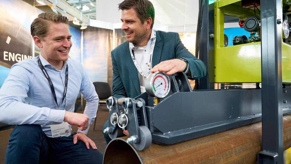 Zwei Männer in Geschäftskleidung untersuchen auf einer Industriemesse ein großes Rohr mit einem Druckmesser.